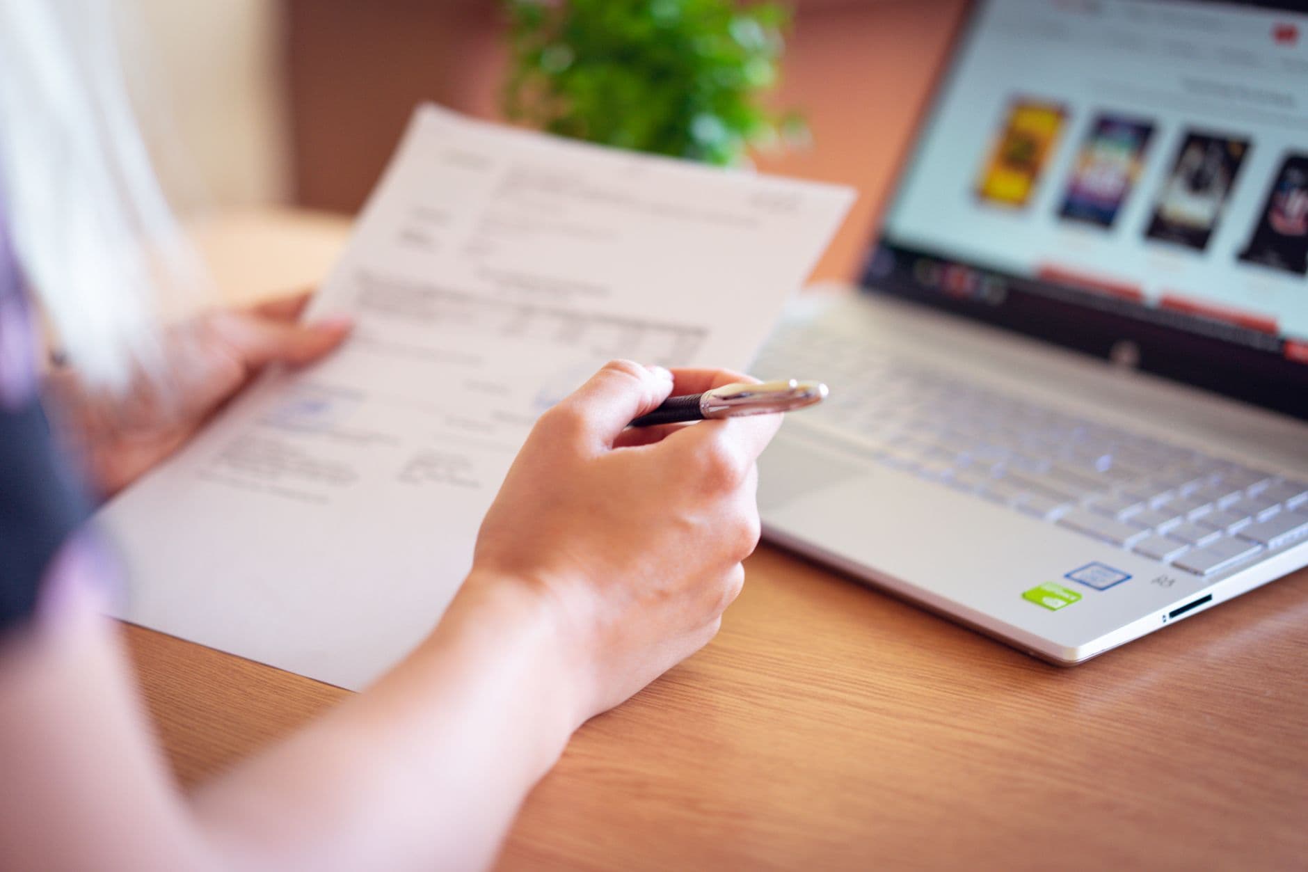 Close-up of a hand holding a pen and paper near a laptop, suggesting online shopping or document review.