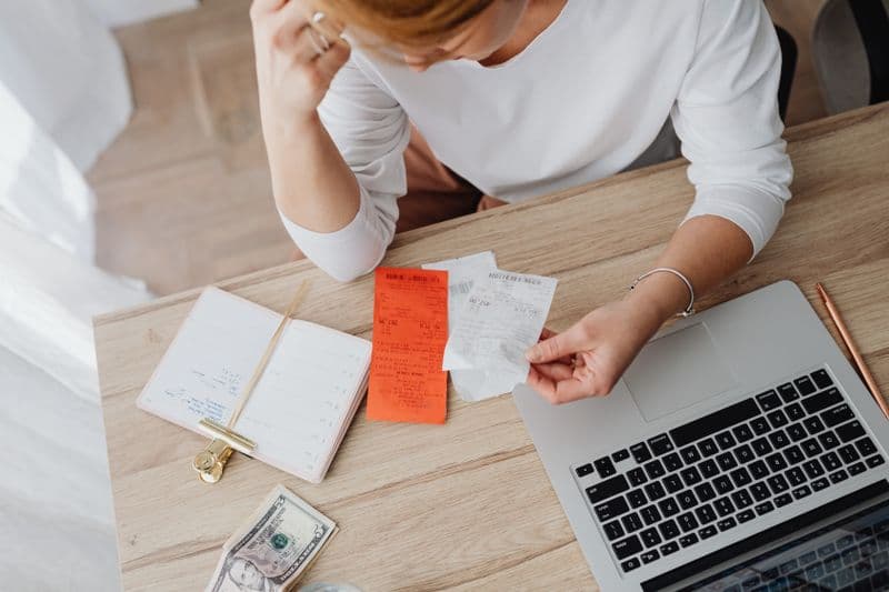 Declaração anual do MEI: como preencher e regularizar Woman reviewing receipts and planning budget using a laptop and notebook at home to manage expenses.