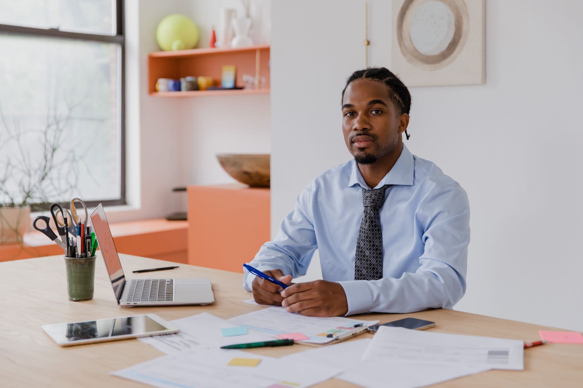 MEI emitindo nota para empresa: o que informar A professional businessman with a beard and braided hair sits at his desk with a laptop and documents, working diligently.