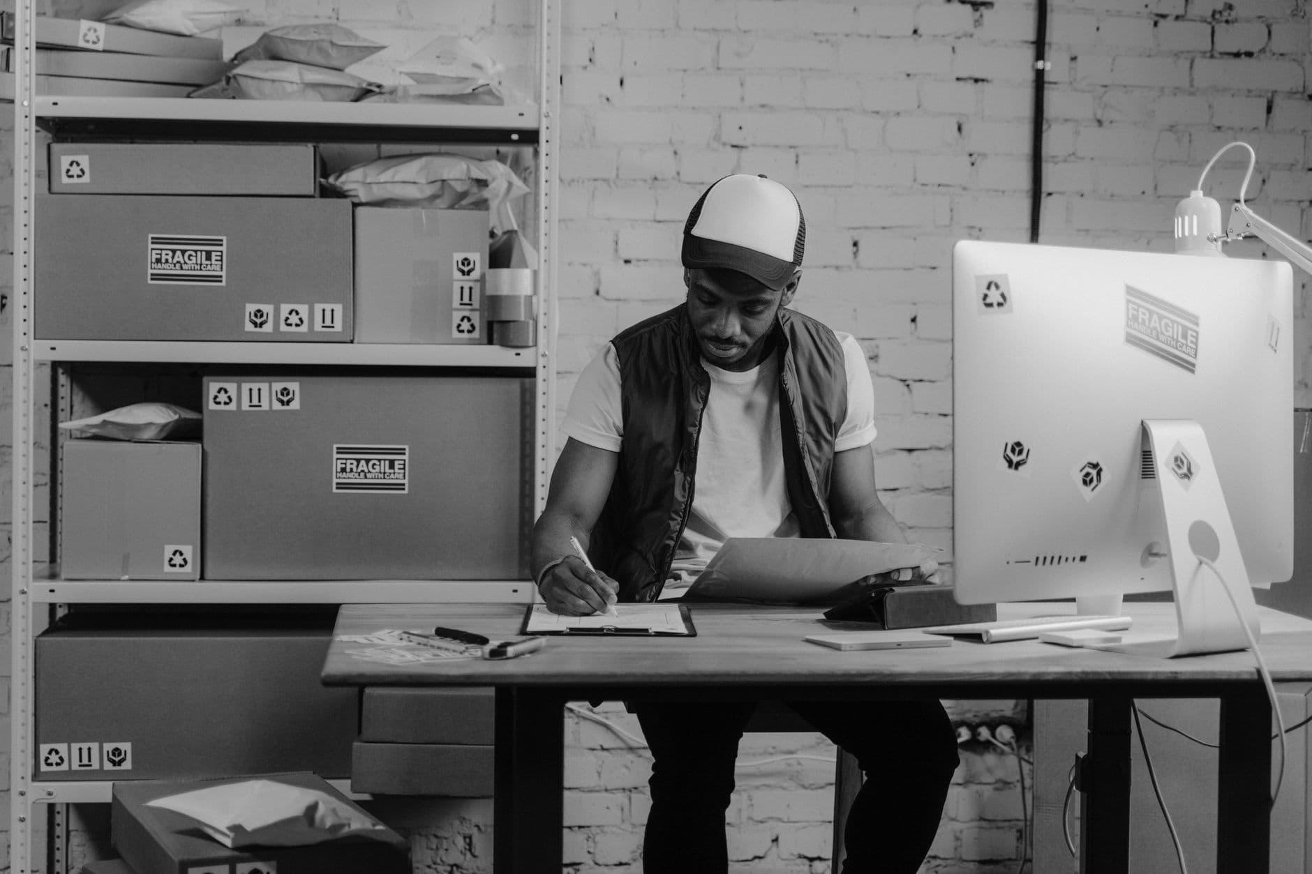 Emissor de NFE gratuita: opções e limitações Warehouse worker organizing shipments at a desk, surrounded by packages and a computer, illustrating logistics work.