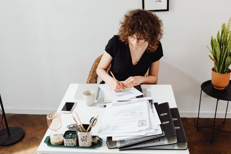 Emitir a nota antes ou depois de receber? Caucasian woman writing at a home office desk, focusing on documents and organization.