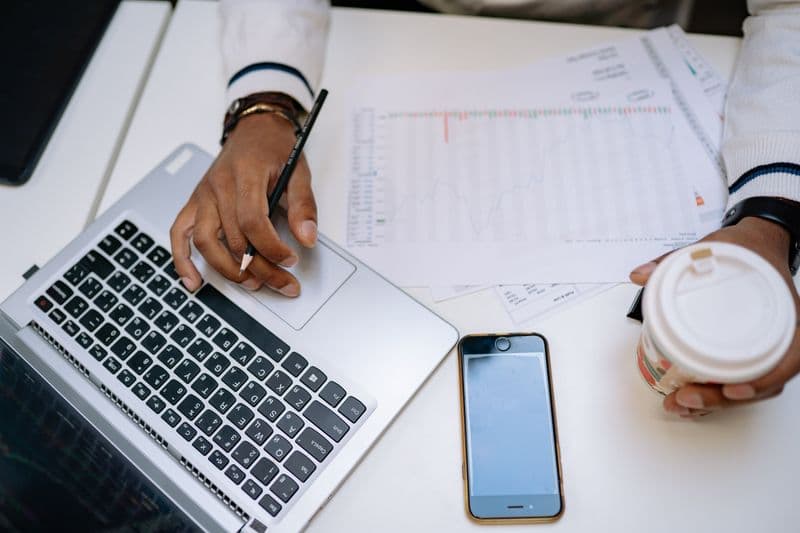 Sua atividade pode ser MEI? Como conferir A businessman holding coffee and using a laptop at a desk with charts and a smartphone.