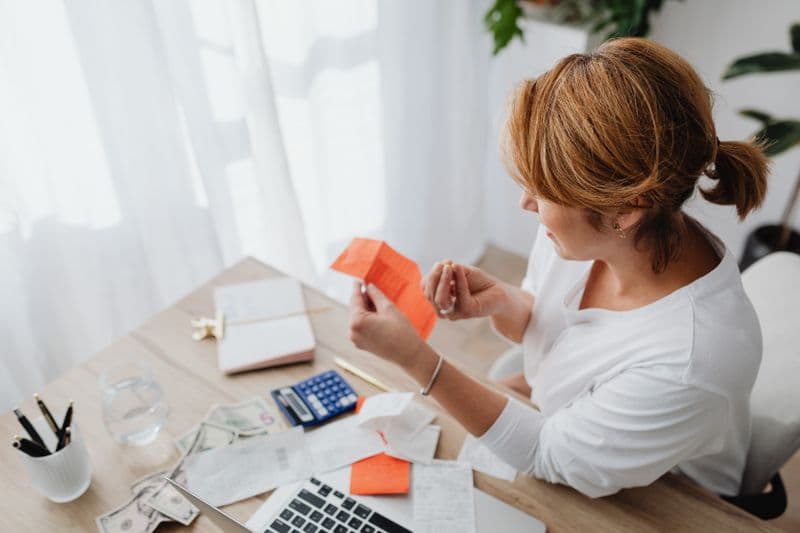 MEI: como declarar lucro isento e pró-labore no IRPF A woman organizing her finances with bills and receipts at a home office desk using a calculator and laptop.