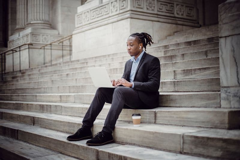 O MEI paga INSS? Entenda os direitos do DAS Full body serious African American businessman with earbuds wearing formal suit sitting on stone stairs of building and browsing modern netbook