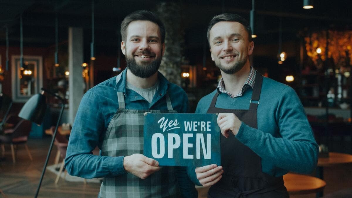 Dois pequenos empreendedores sorridentes segurando placa de aberto na porta do negócio