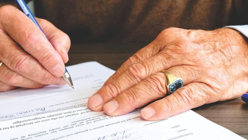 Mão de pessoa idosa assinando um contrato sobre a mesa