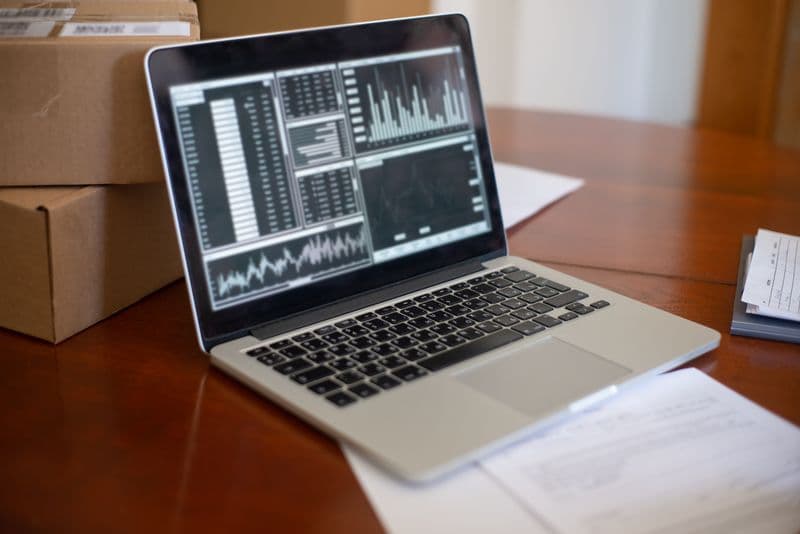 MEI pode ter mais de uma atividade? Veja como incluir Laptop showing data graphs on wooden table with cardboard boxes, depicting a small business office setting.