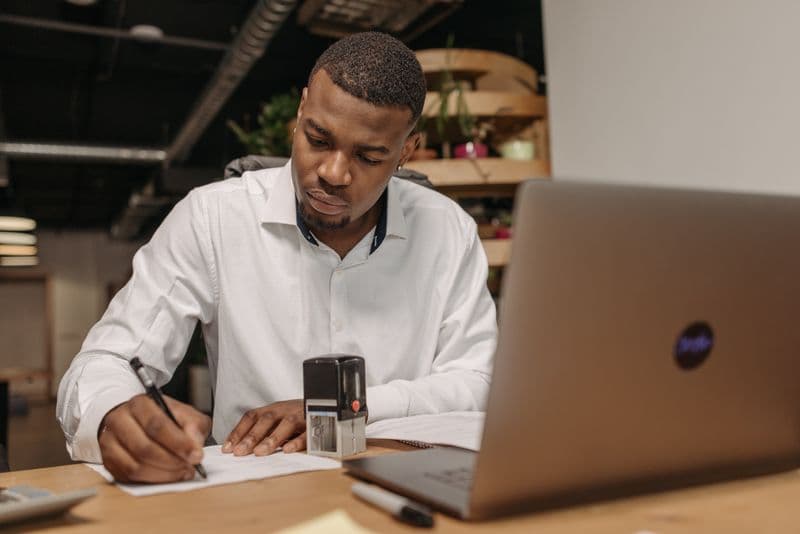 MEI vendendo online: quem emite a nota fiscal? A young man in a white shirt works diligently at his office desk with a laptop and documents.