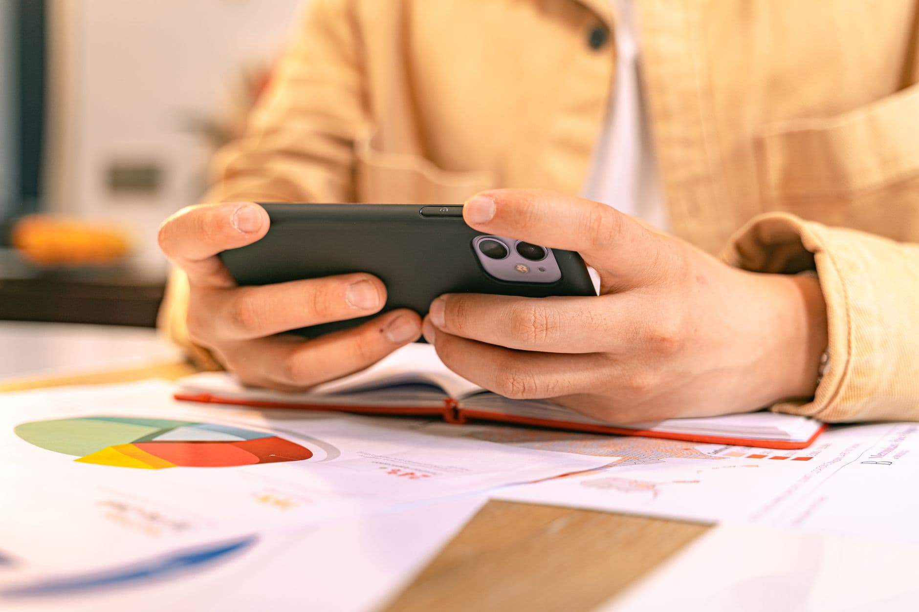 Emitir NFS-e pelo celular: aplicativos oficiais A business person checking their phone while working with graphs and charts on a desk.