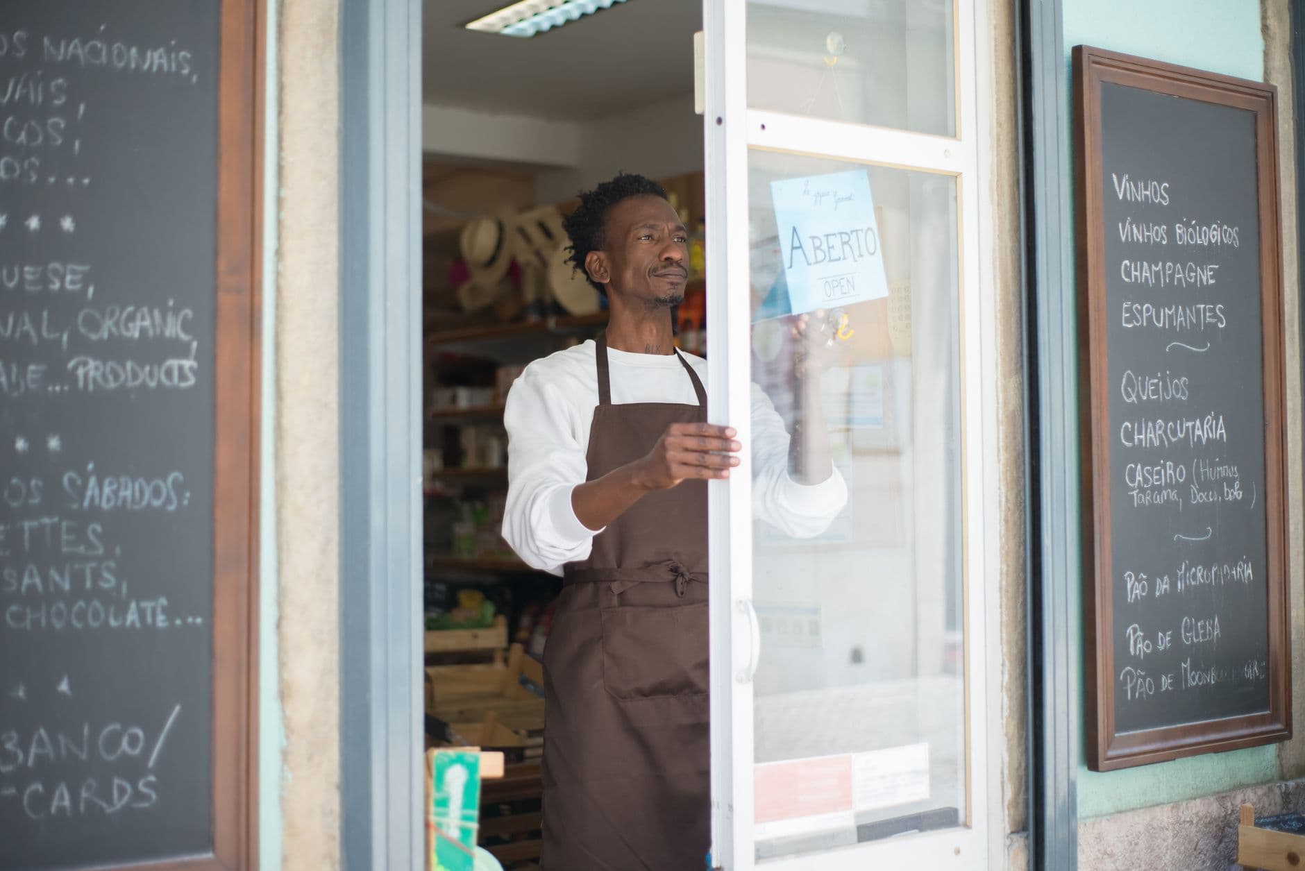 Como emitir NFS-e em Campo Grande A man in an apron opens a grocery store door in Portugal, ready for business.