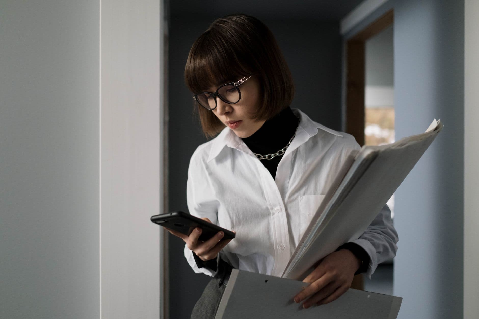 Para que serve o QR Code na nota fiscal Young woman with eyeglasses, holding documents, checking smartphone indoors.