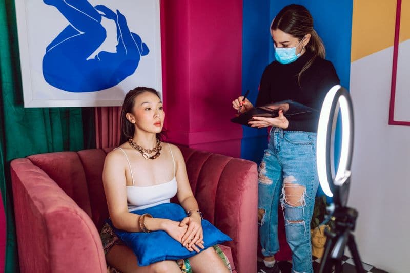Nota fiscal para cabeleireiro autônomo e salão parceiro A makeup artist wearing a face mask applies makeup to a woman in a colorful room.