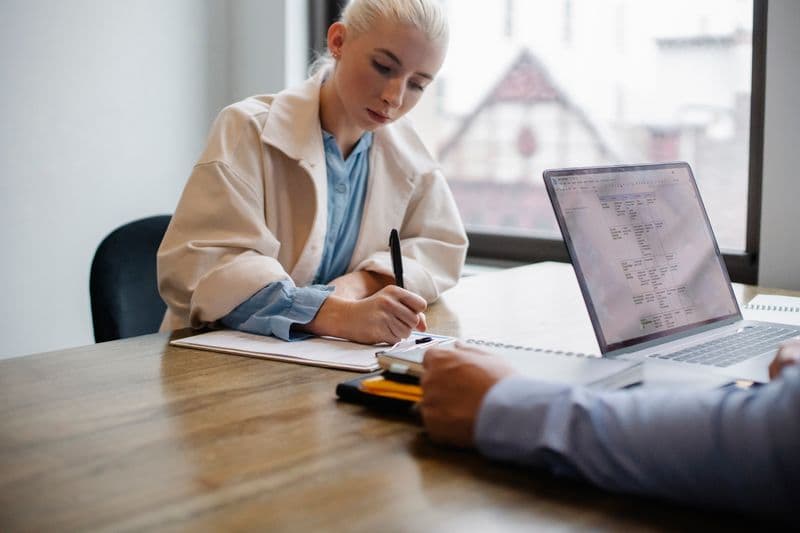 Nota fiscal para coach, mentor e consultor: como emitir Serious young female manager in formal outfit sitting and signing documents while business partner working on laptop