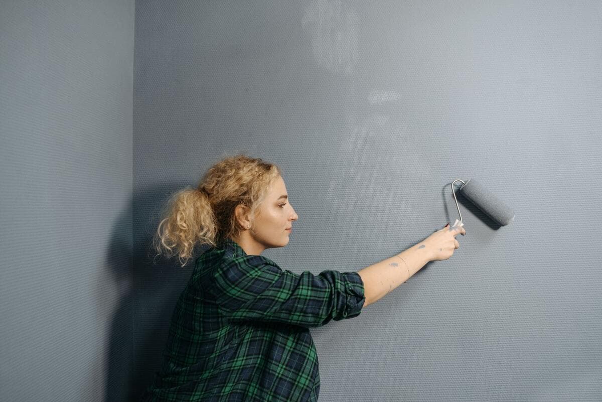 O material da obra e onde pagar o ISS Mulher pintando uma parede de cinza com rolo de pintura