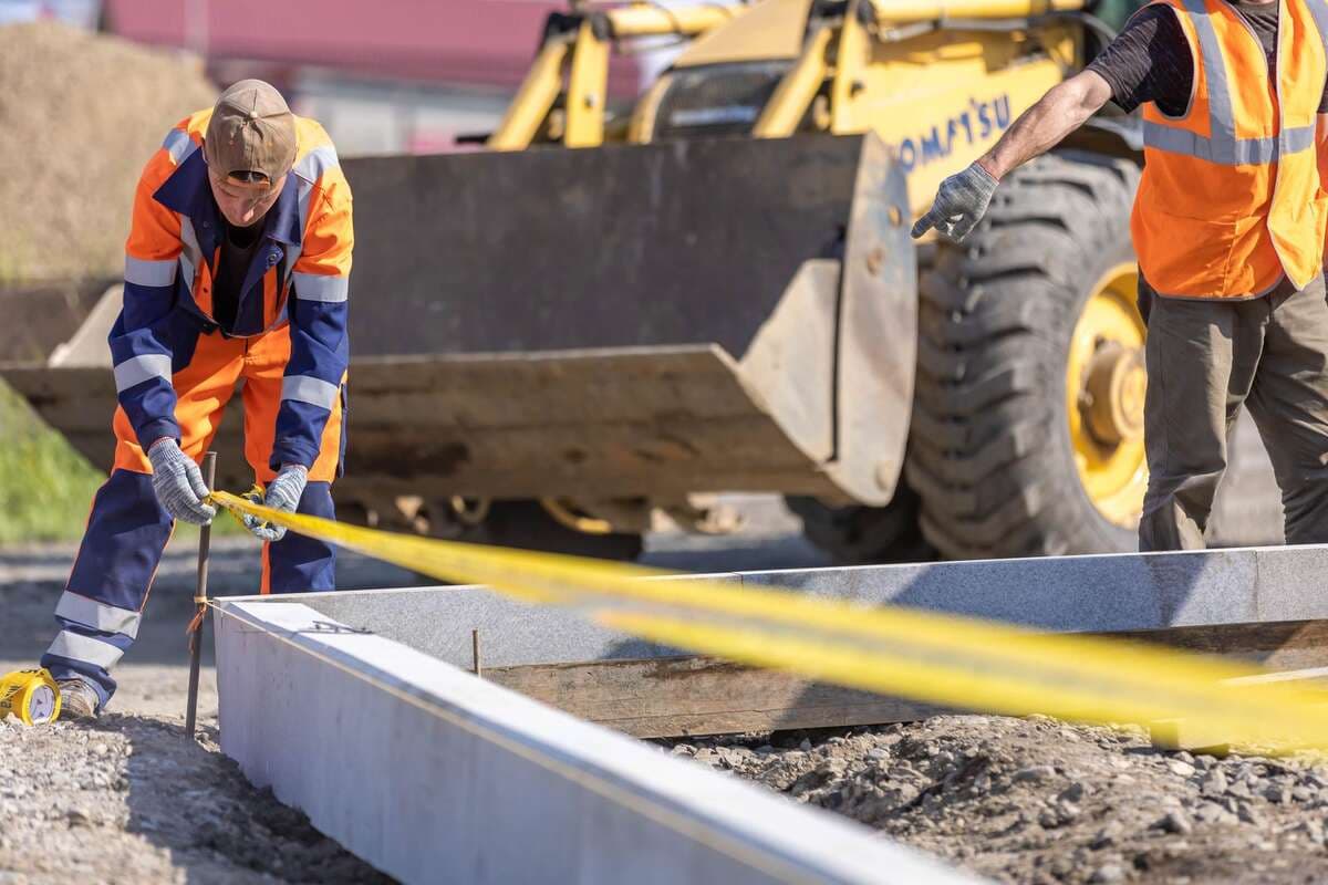Nota fiscal ajuda a conseguir mais obras Trabalhadores medindo com trena em uma obra com escavadeira ao fundo