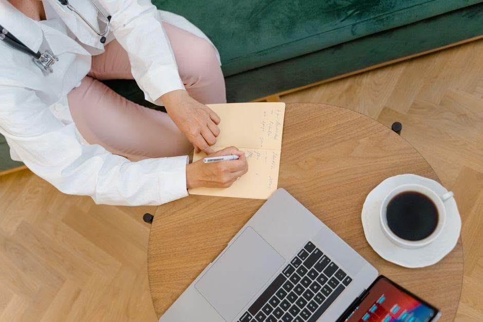 Profissional de saúde fazendo anotações em consulta, com notebook e café na mesa