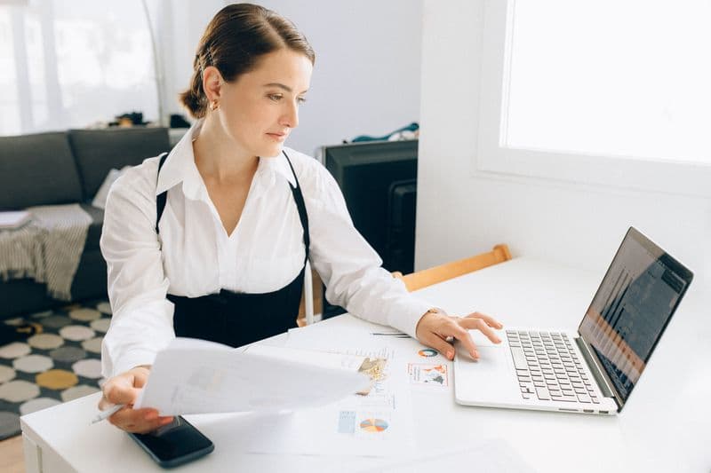 Separar as contas pessoal e da empresa: por onde começar Woman at desk reviewing business reports on laptop, holding papers in a modern office setting.